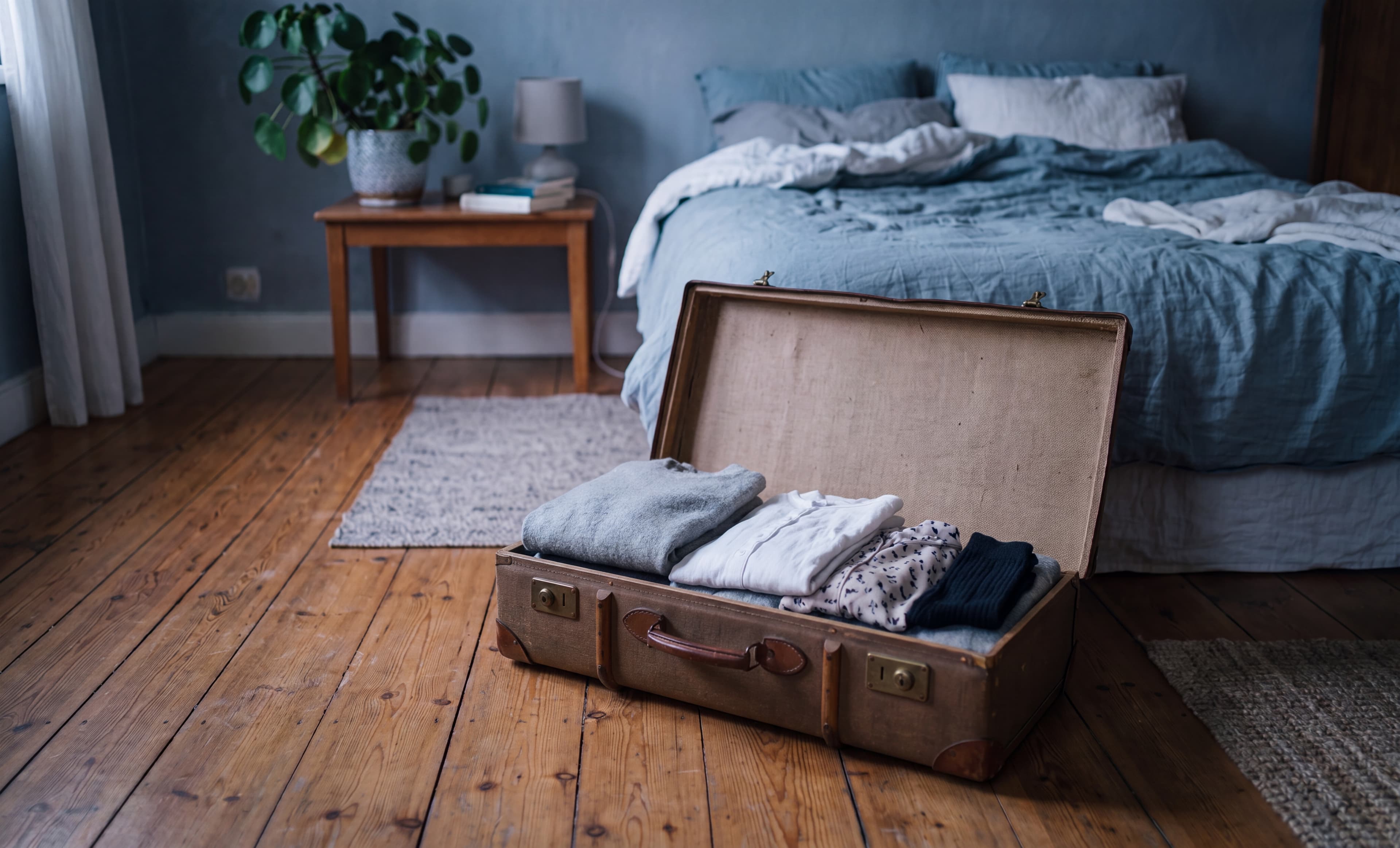 Ein halb gepackter Vintage-Koffer auf Dielenboden vor einem blau gestrichenen Schlafzimmer im Dämmerlicht.